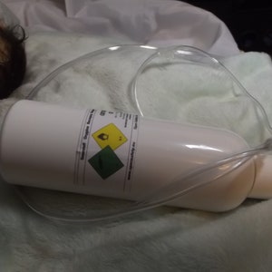 May include: A white oxygen tank with a yellow and green warning label. The tank is connected to a clear plastic tube.