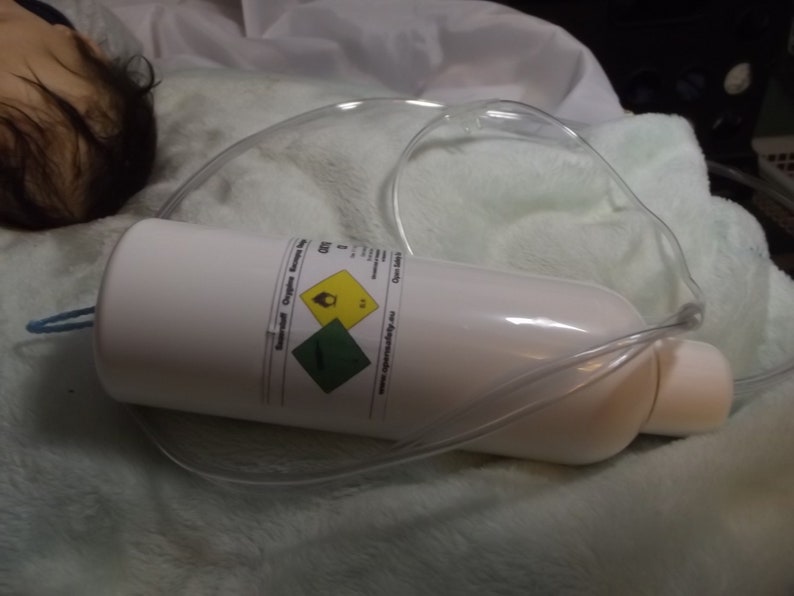 May include: A white oxygen tank with a yellow and green warning label. The tank is connected to a clear plastic tube.