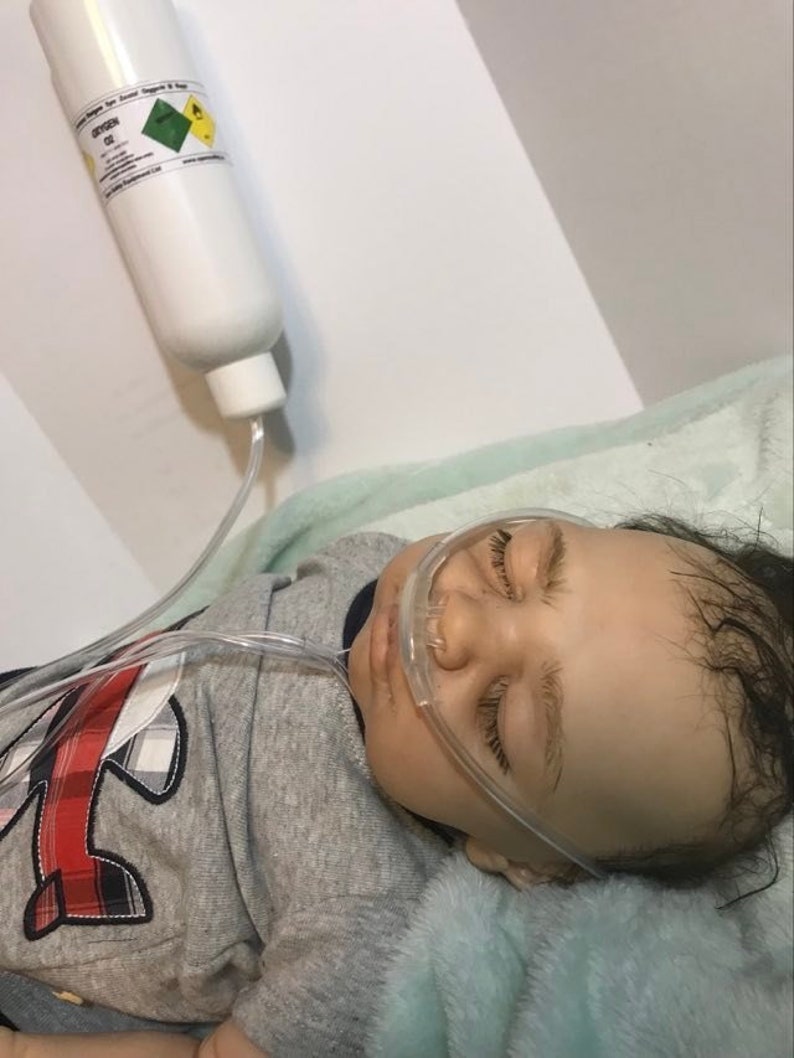 May include: A baby doll with brown hair and closed eyes is lying on a light blue blanket. The doll is wearing a gray shirt with a red airplane design. A clear plastic tube is connected to the doll's nose and a white bottle labeled "Oxygen O2" is hanging above the doll.