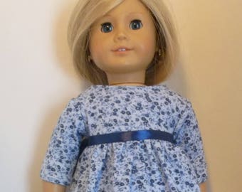 May include: A doll wearing a light blue dress with a floral pattern and a dark blue ribbon belt. The doll has blonde hair and is wearing black leggings. The dress has three-quarter sleeves.