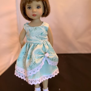 May include: A doll with brown hair wearing a light blue dress with a floral pattern and white lace trim. The doll is wearing white shoes and socks. The dress features a bow on the front.
