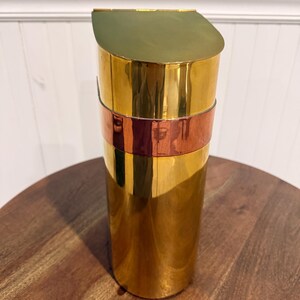 May include: A tall, cylindrical container with a gold-colored body and a copper-colored band. The top is a gold-colored, angled lid. The container is sitting on a wooden surface.