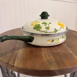 May include: Cream-colored enamel saucepan with a matching lid. The pan and lid feature a floral design in yellow and green. The handle and lid knob are dark green. The pan is sitting on a wooden table.