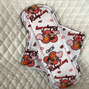 Puede incluir: Compresa menstrual de tela reutilizable con fondo blanco y un estampado con una pareja de dibujos animados, corazones y el texto "Ricky Lucy" y "XOXO". La compresa tiene un reverso azul oscuro y un cierre de botón a presión rojo.