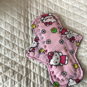 May include: A pink reusable menstrual pad featuring a Hello Kitty design. The pad has a pink base with white Hello Kitty characters, flowers, and other small graphics. The pad is edged with black trim and has a snap closure.