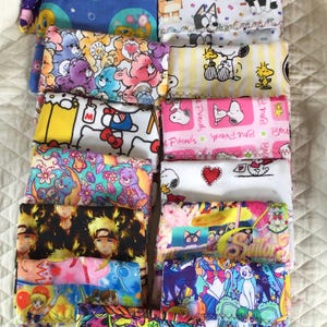 Puede incluir: Una colección de bolsas de tela coloridas, cada una con un diseño diferente de personaje de dibujos animados o anime. Los diseños incluyen My Little Pony, Care Bears, Snoopy, Hello Kitty y Sailor Moon. Las bolsas están apiladas.