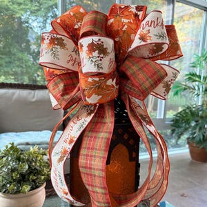 May include: A black metal lantern topped with a large decorative bow made of wired ribbons. The ribbons feature autumn leaf patterns in orange, red, and yellow, a green and orange plaid design, and white fabric with the printed text Thankful.