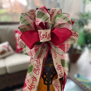 Puede incluir: Un lazo de cinta a cuadros rojo y verde con el texto "Merry Christmas" y "Merry Christmas" en él. El lazo está atado alrededor de una linterna negra con un panel de vidrio transparente.