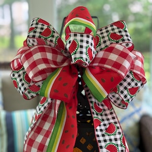 May include: A decorative bow featuring a watermelon theme. The bow is made of red and white gingham, black and white checkered, and watermelon-printed ribbons. Green and yellow striped ribbon accents the design. The bow is likely used for seasonal decor.