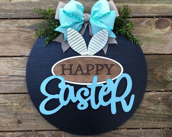May include: A black round wooden door hanger with the words "Happy Easter" in blue lettering. A blue and white polka dot bow with bunny ears is attached to the top.