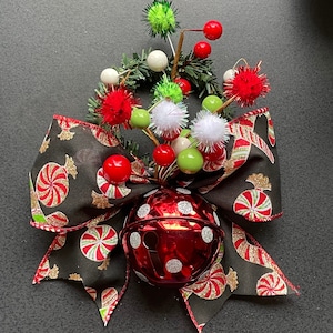 Może przedstawiać: Czerwona ozdoba w kształcie dzwonka w białe kropki z czarną i białą wstążką w kształcie cukierka. Wstążka jest ozdobiona złotym brokatem i zielonymi i czerwonymi jagodami. Ozdoba jest otoczona zielonymi i białymi pomponami.