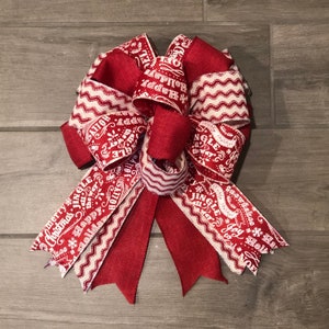 May include: A red and white Christmas bow with a burlap center. The bow is made of two different ribbons, one with a white chevron pattern and the other with white text on a red background. The text on the ribbon includes "Jingle All The Way", "Merry Christmas", "Happy Holidays", "Snowflakes", "Joy", and "Holly".