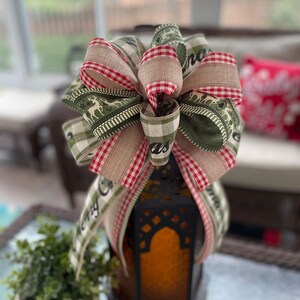 Puede incluir: Un lazo de cuadros verdes y rojos con una cinta de "Feliz Navidad" atada a una linterna negra. El lazo tiene un patrón de cuadros blancos y verdes con un diseño de reno.