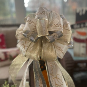 May include: A decorative Christmas bow featuring layers of white, gold, and gray ribbons. The ribbons are adorned with gold text that reads "Christmas". The bow is designed with a layered, full shape, perfect for holiday decor.