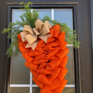 May include: Orange and brown burlap carrot shaped door wreath with a brown burlap bow and green sprigs.