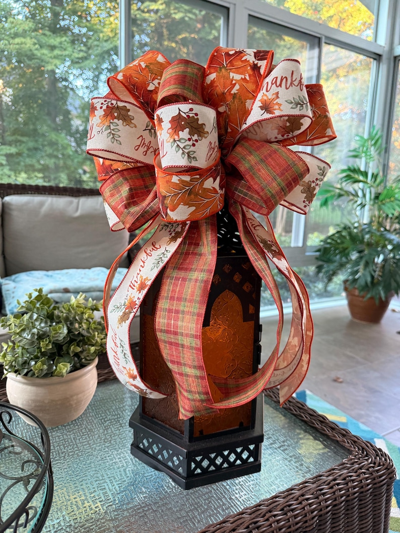 May include: A black metal lantern topped with a large decorative bow made of wired ribbons. The ribbons feature autumn leaf patterns in orange, red, and yellow, a green and orange plaid design, and white fabric with the printed text Thankful.