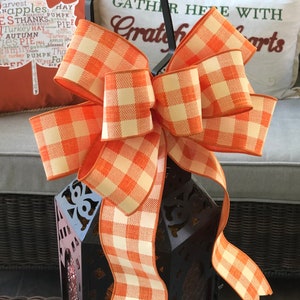 May include: A large orange and white checkered bow tied around a black metal lantern. The lantern has a decorative design and is sitting on a table. The bow is made of a soft, textured fabric.