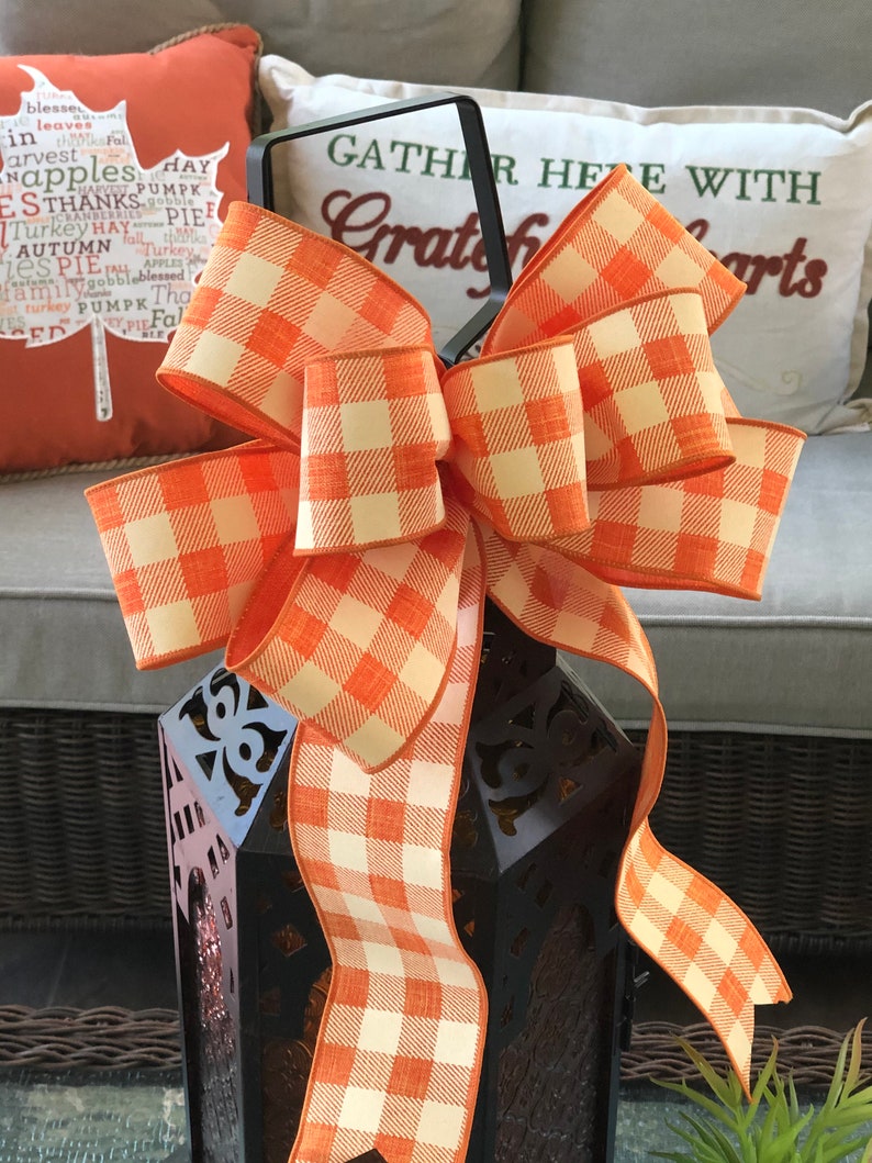 May include: A large orange and white checkered bow tied around a black metal lantern. The lantern has a decorative design and is sitting on a table. The bow is made of a soft, textured fabric.