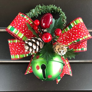May include: A green bell with red polka dots is surrounded by a red and green bow with white polka dots. The bow is decorated with pine cones and red berries.