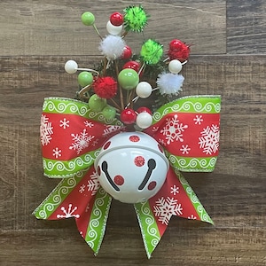 以下が含まれることがあります： 中央に白いベルのオーナメントが付いた赤と緑のクリスマスリボン。リボンは白い雪片と緑色の渦巻きで飾られています。ベルには赤い水玉模様があります。