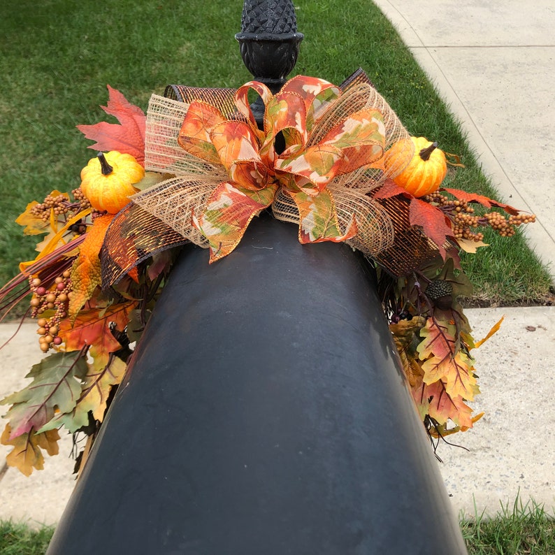 May include: A black lamp post adorned with a fall-themed arrangement. The arrangement includes a large bow with orange, brown, and green accents, small pumpkins, and colorful fall leaves. The overall design evokes a seasonal, autumnal aesthetic.