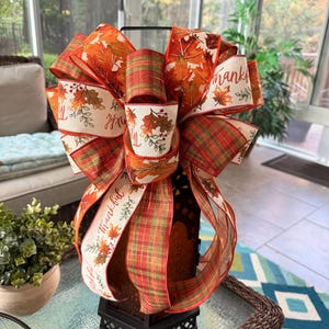 May include: A decorative bow featuring a mix of ribbons with autumn leaf patterns in orange, red, and yellow, alongside a green and red plaid ribbon. The cream-colored ribbon includes the printed text Thankful with small leaf and berry accents.