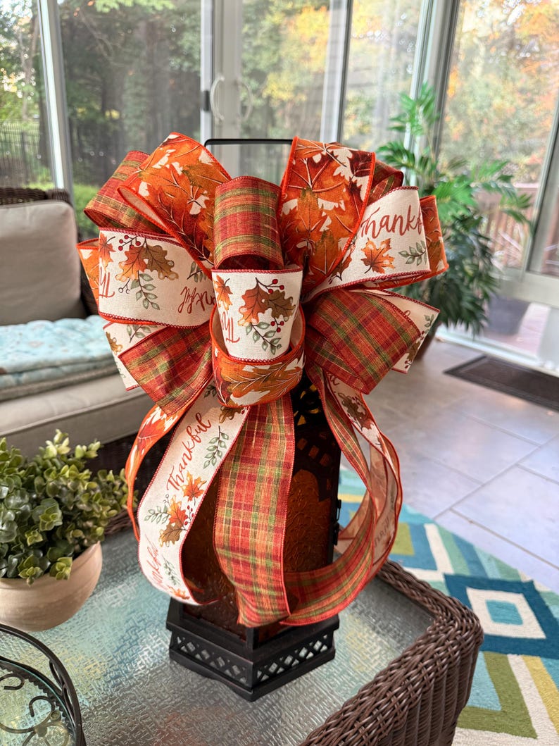 May include: A decorative bow featuring a mix of ribbons with autumn leaf patterns in orange, red, and yellow, alongside a green and red plaid ribbon. The cream-colored ribbon includes the printed text Thankful with small leaf and berry accents.