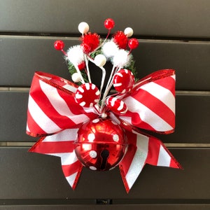 Pode incluir: Um laço listrado vermelho e branco com um sino vermelho e enfeites de bastão de caramelo branco e vermelho. O laço é decorado com glitter branco.