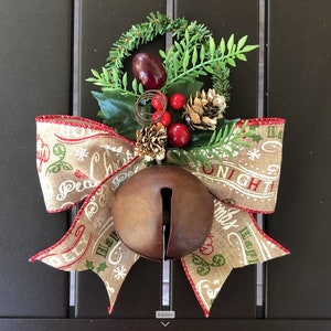 以下が含まれることがあります： 赤と緑のクリスマスの飾りで飾られた、麻ひもで作ったリボンが付いた、素朴な茶色の金属製のベル。リボンには「Peace on Earth」と「Happy Holidays」の文字が印刷されています。