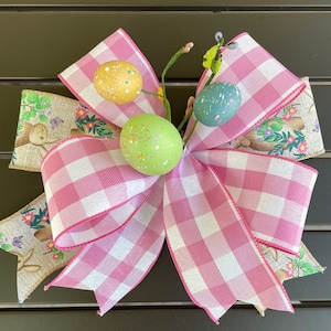 May include: A pink and white gingham bow with a brown burlap ribbon and three pastel Easter eggs. The eggs are green, yellow, and blue.