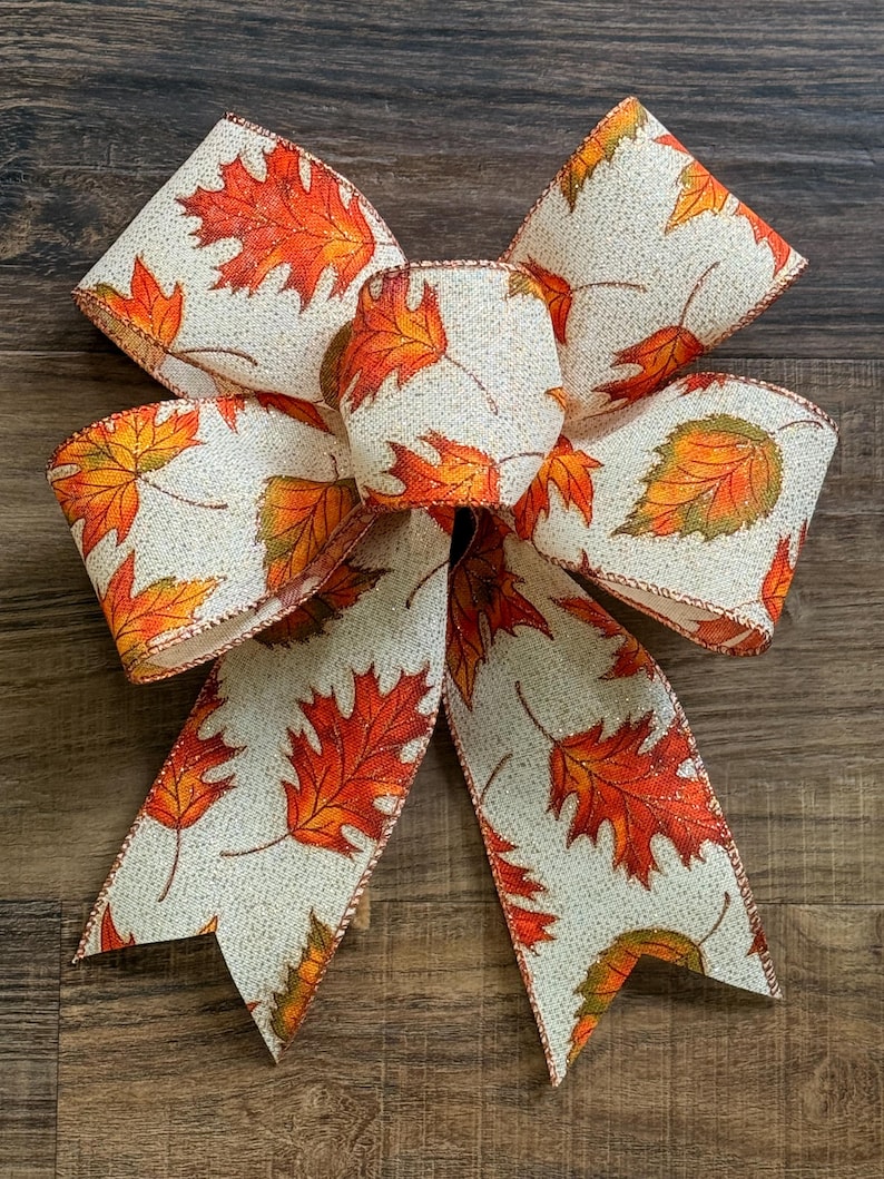 May include: A decorative bow made of cream-colored fabric featuring a repeating pattern of orange, red, and green autumn leaves. The bow has two large upper loops, a central knot, and two long, downward-hanging tails with V-shaped notched ends.