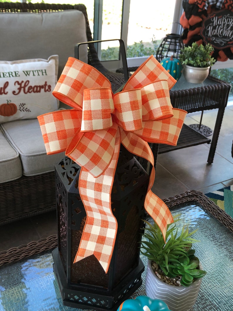 May include: A black metal lantern with an orange and white checkered bow. The lantern has a decorative design and is sitting on a table.