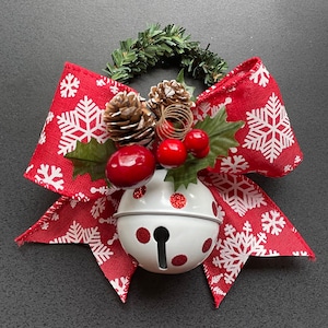 May include: A red and white bow with a white snowflake pattern and a white bell with red polka dots. The bow is decorated with a pine cone, red berries, and green leaves.