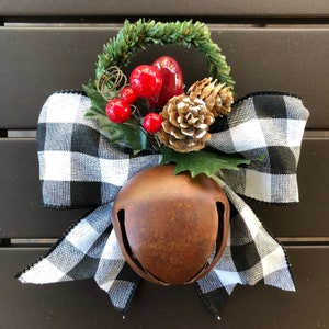 May include: A rustic brown metal bell with a black and white buffalo plaid bow and a green sprig of holly with red berries and a pine cone.