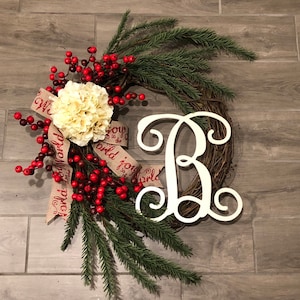 May include: A Christmas wreath with a grapevine base, adorned with faux red berries, evergreen sprigs, and a cream-colored flower. A burlap ribbon with the words "Joy to the World" and a white letter "B" complete the festive design.