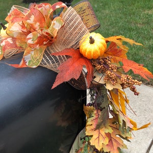 May include: A black mailbox decorated with fall-themed accents. The arrangement includes a large bow with orange and gold ribbons, faux pumpkins, and colorful autumn leaves. A decorative acorn finial is visible.