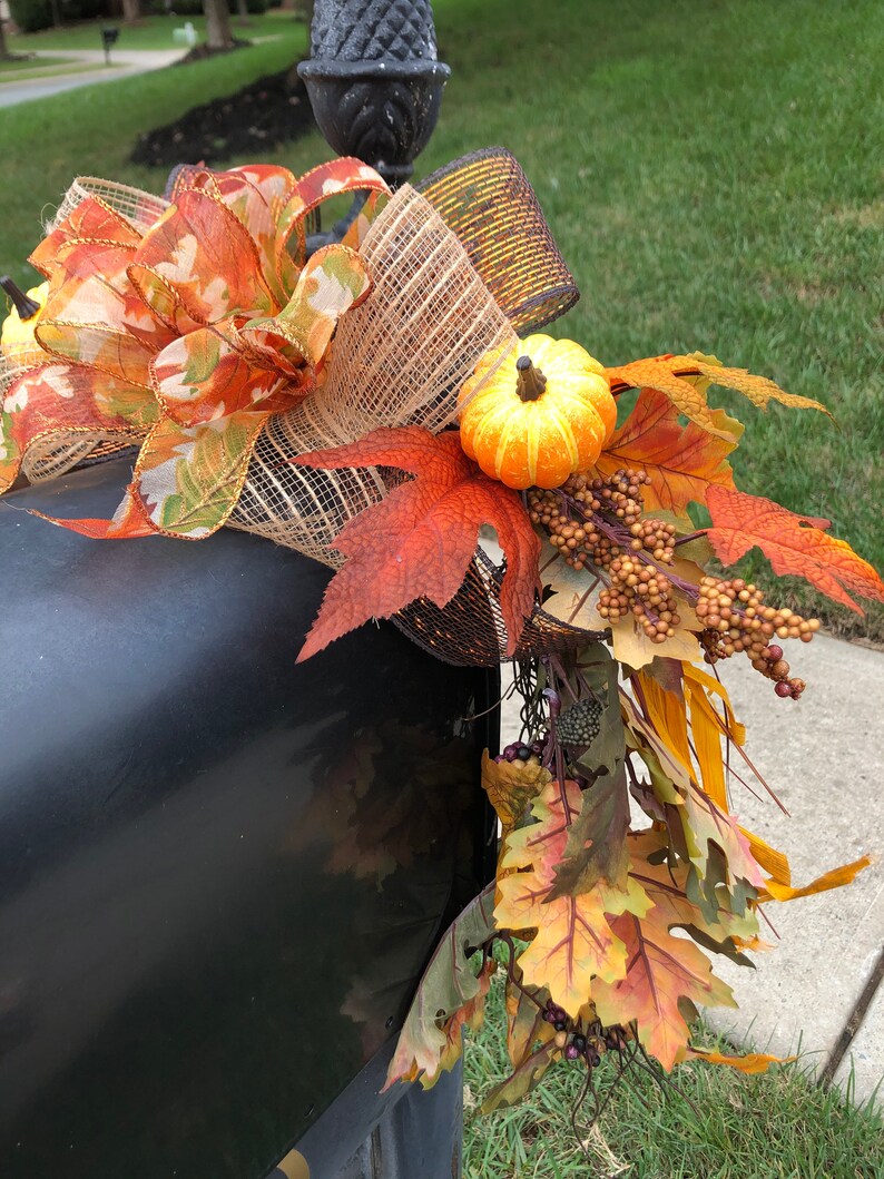 May include: A black mailbox decorated with fall-themed accents. The arrangement includes a large bow with orange and gold ribbons, faux pumpkins, and colorful autumn leaves. A decorative acorn finial is visible.
