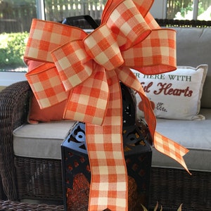 May include: A large orange and white gingham bow tied around a black lantern. The lantern is sitting on a table with a wicker chair and a pillow that says "Gather Here With Grateful Hearts".