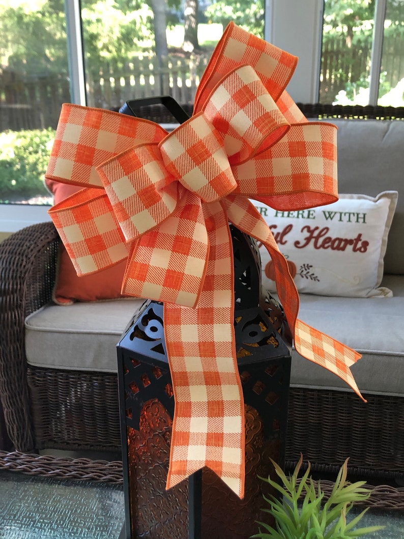 May include: A large orange and white gingham bow tied around a black lantern. The lantern is sitting on a table with a wicker chair and a pillow that says "Gather Here With Grateful Hearts".