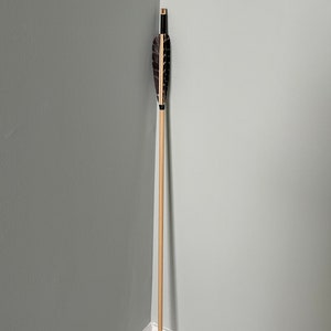 Puede incluir: Una flecha de madera con plumas. Las plumas son negras y marrones con detalles dorados. La flecha está de pie en la esquina de una habitación con paredes grises.