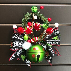 May include: A green jingle bell with red glitter dots, surrounded by a black and white ribbon with the text "Joy to the World" and "Happy Holidays". The bell is decorated with red and green candy canes, white pom-poms, and green and red berries.