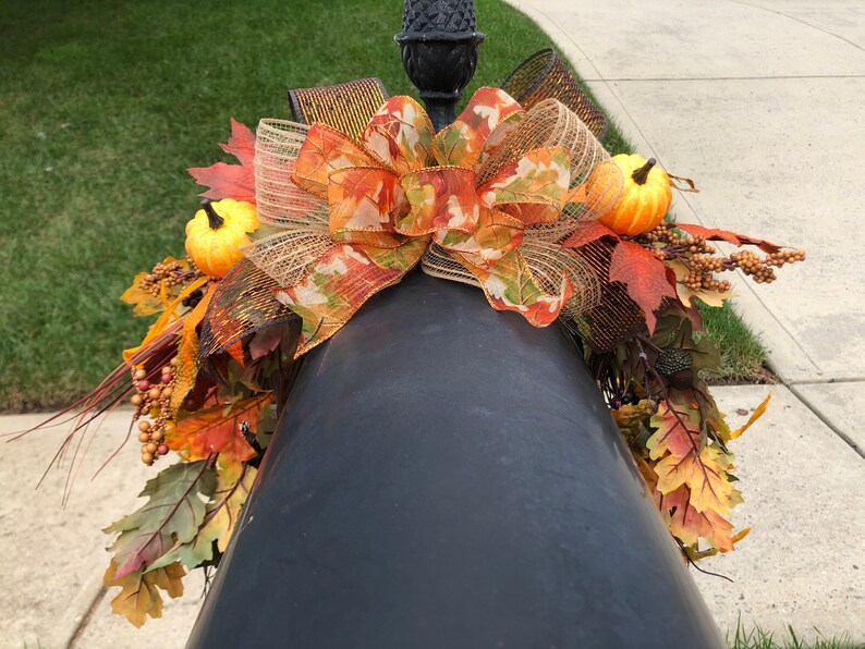May include: A fall-themed decorative arrangement featuring a large bow with orange, green, and brown ribbons, adorned with small pumpkins, fall leaves, and berries. The arrangement is placed on a black post, creating a festive autumn display.