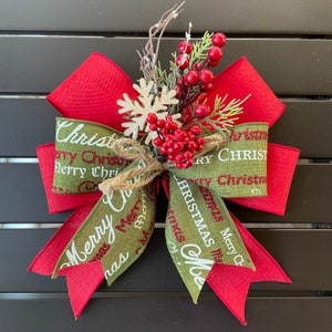 以下が含まれることがあります： 赤いリボンと緑のリボンでできたクリスマスの飾り。麻ひもで結ばれており、赤い実と白い雪の結晶がついた緑の葉が飾られています。リボンには「Merry Christmas」と「Christmas」の文字が白く書かれています。