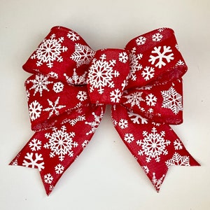 May include: A red bow with white snowflake patterns. The bow is made of fabric and is tied in a classic bow shape.