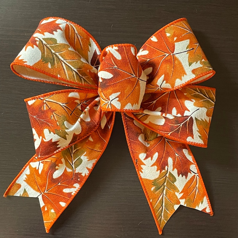 May include: A large, orange and white bow with a fall leaf print. The bow is made of fabric and has a wired edge.
