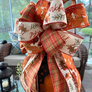 May include: A decorative bow made of wired ribbon featuring a fall theme with orange, red, and green plaid patterns, autumn leaves, and berries. The cream-colored ribbon sections are printed with the word thankful in a cursive script font.