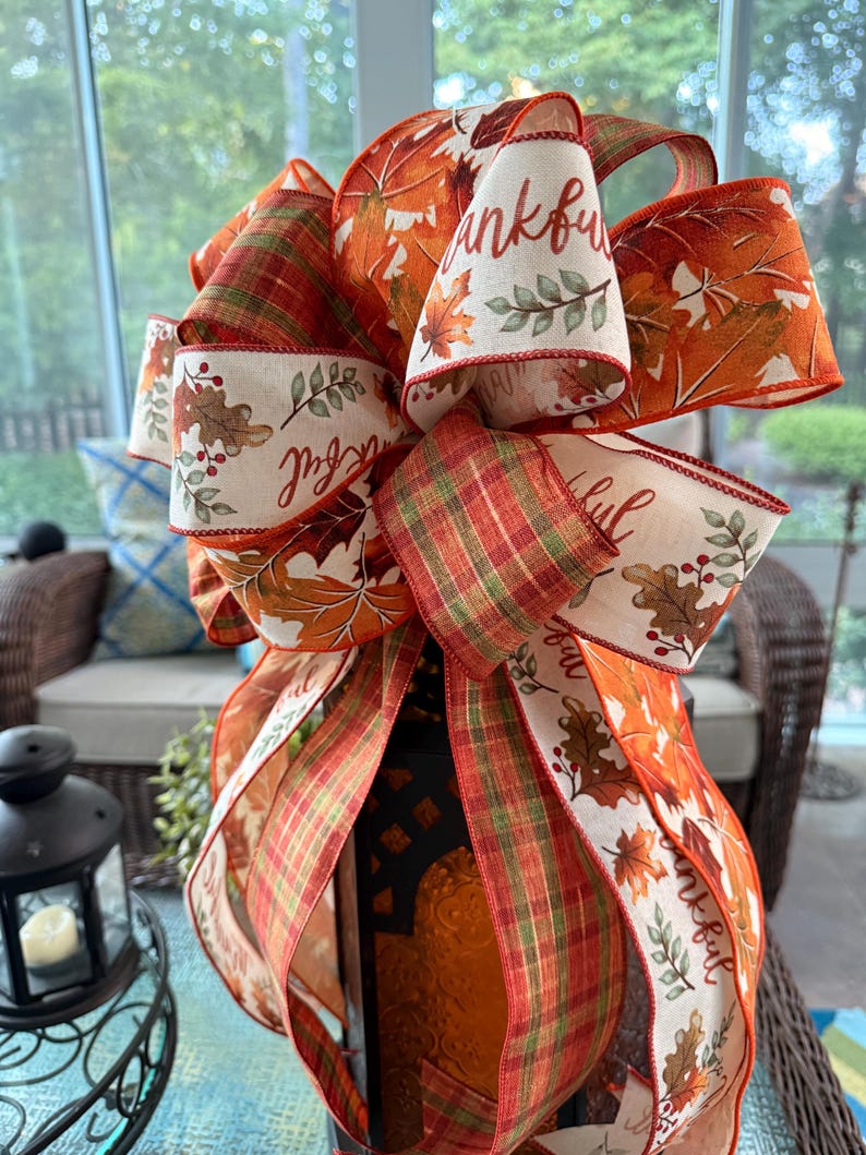 May include: A decorative bow made of wired ribbon featuring a fall theme with orange, red, and green plaid patterns, autumn leaves, and berries. The cream-colored ribbon sections are printed with the word thankful in a cursive script font.