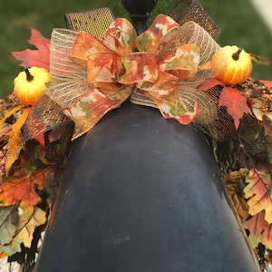 May include: A fall-themed decorative arrangement featuring a large bow with orange, brown, and gold ribbons, accented with small pumpkins, fall leaves, and berries. The arrangement is placed on a dark, possibly metallic, surface.