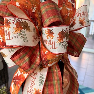 May include: A decorative bow made of wired ribbon featuring a pattern of autumn leaves in shades of orange, red, and brown, interspersed with cream-colored sections printed with the word Thankful and small leaf illustrations, plus plaid ribbon.
