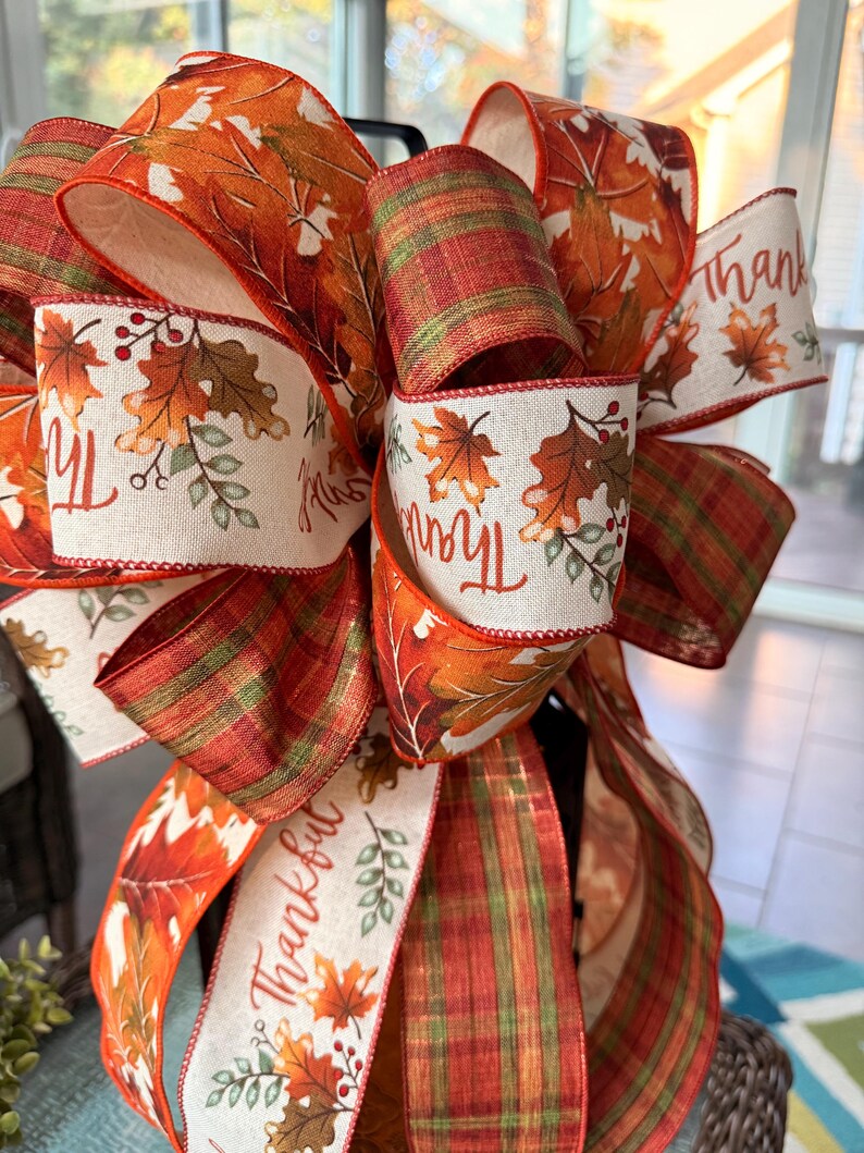 May include: A decorative bow made of wired ribbon featuring a pattern of autumn leaves in shades of orange, red, and brown, interspersed with cream-colored sections printed with the word Thankful and small leaf illustrations, plus plaid ribbon.